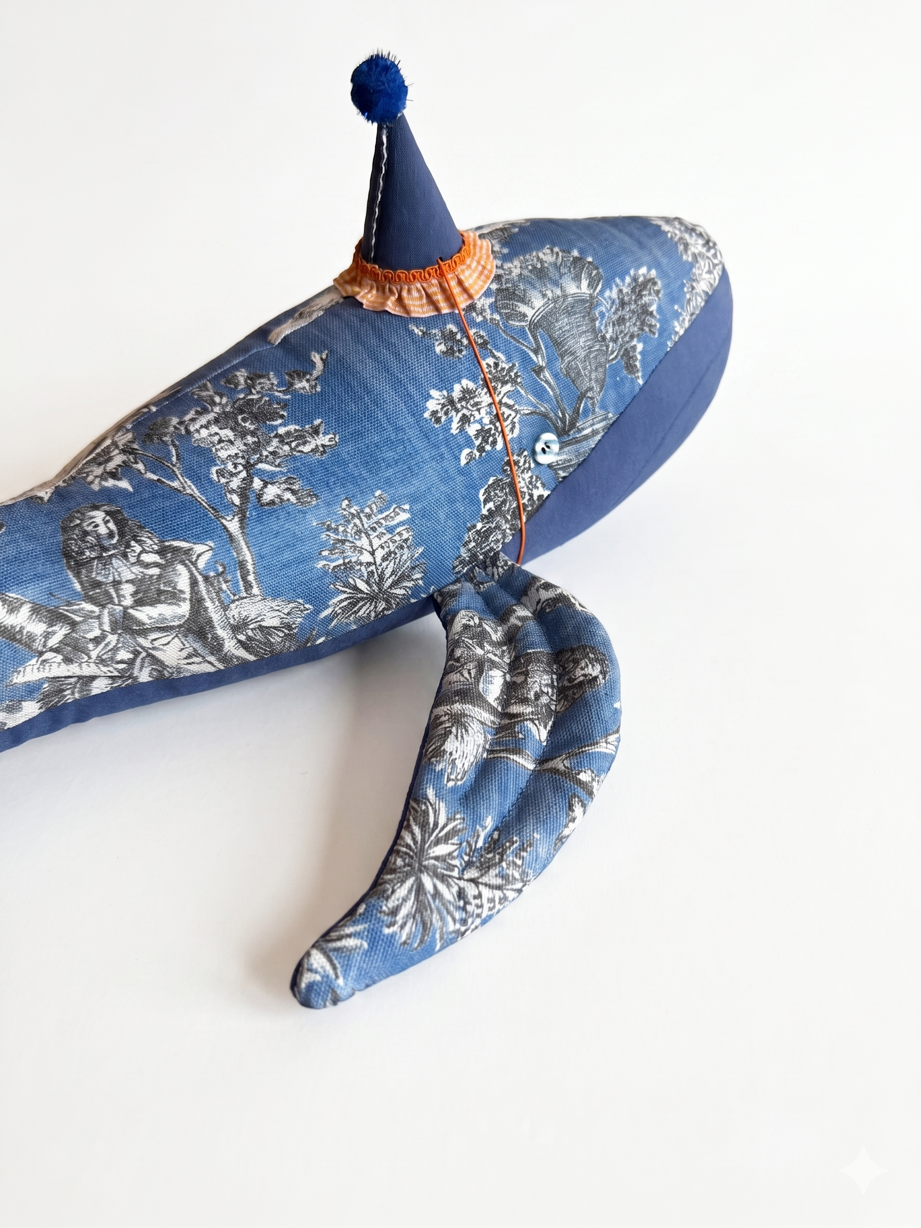 Giocattolo balena con cappuccio blu e ponpon, bavetta arancione, stampa grigia.