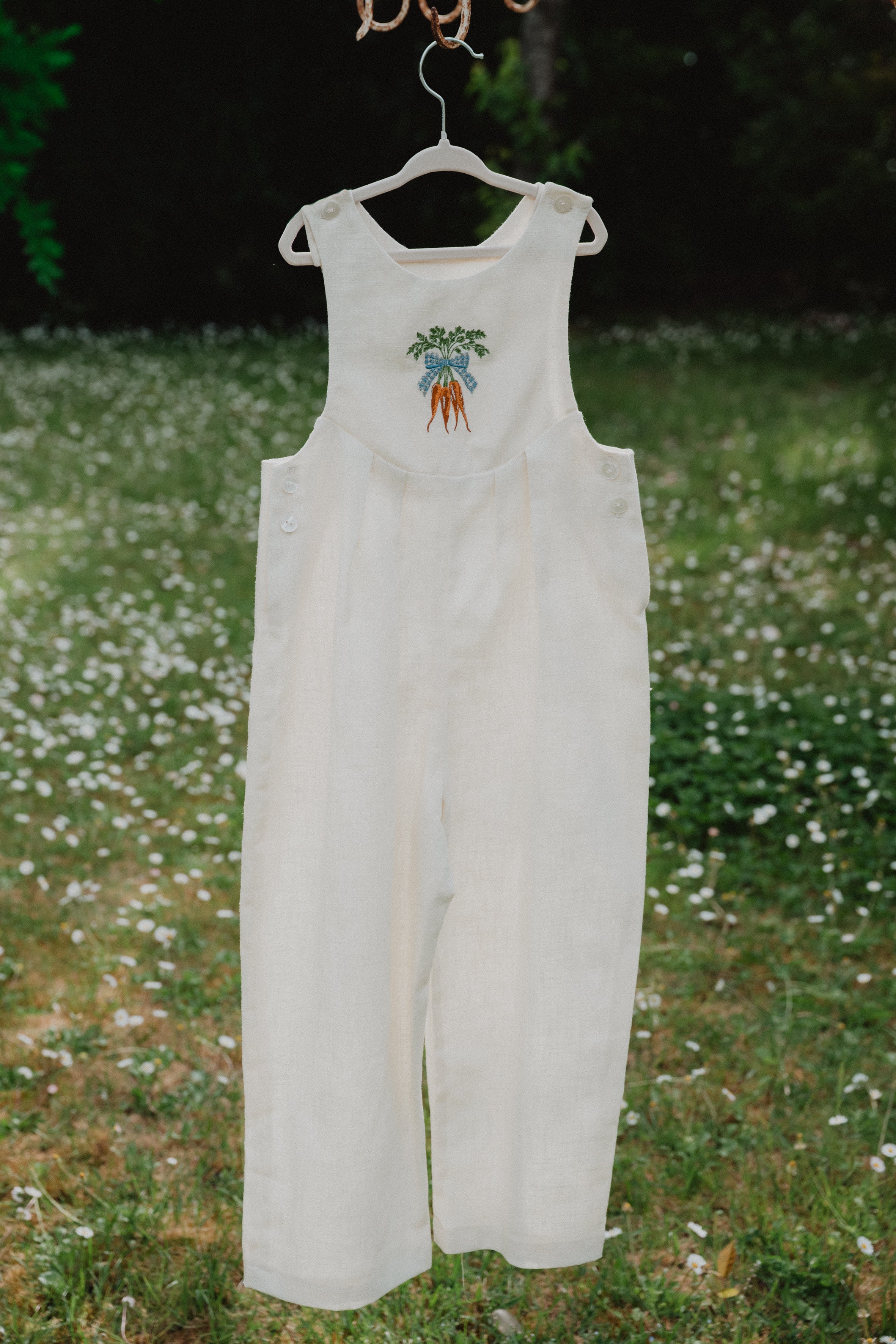 Salopette unisex in lino lavato bianco panna appesa, con ricamo carote sulla pettorina