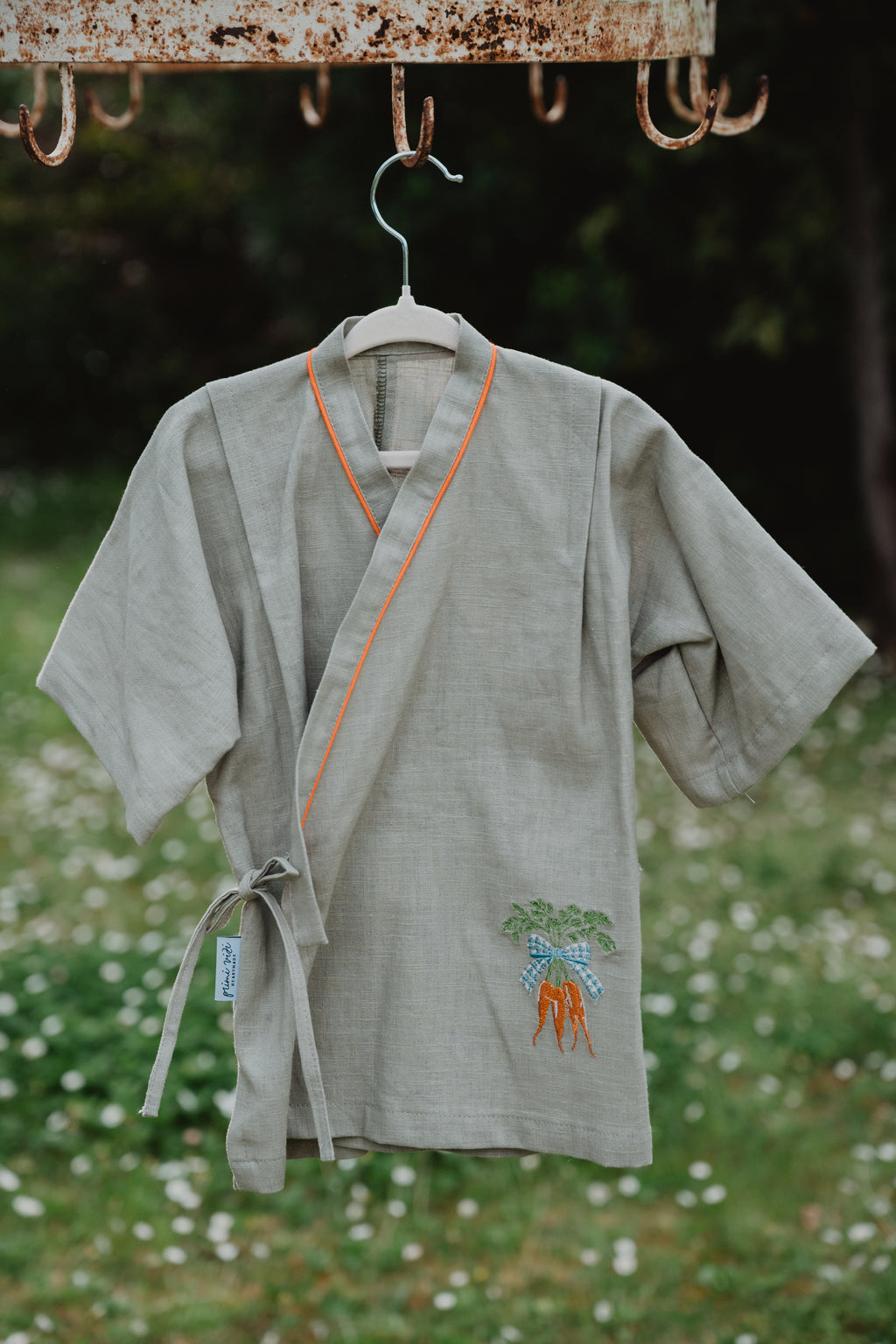 Camicia kimono in lino lavato verde oliva appesa, bordo arancione, ricamo carote