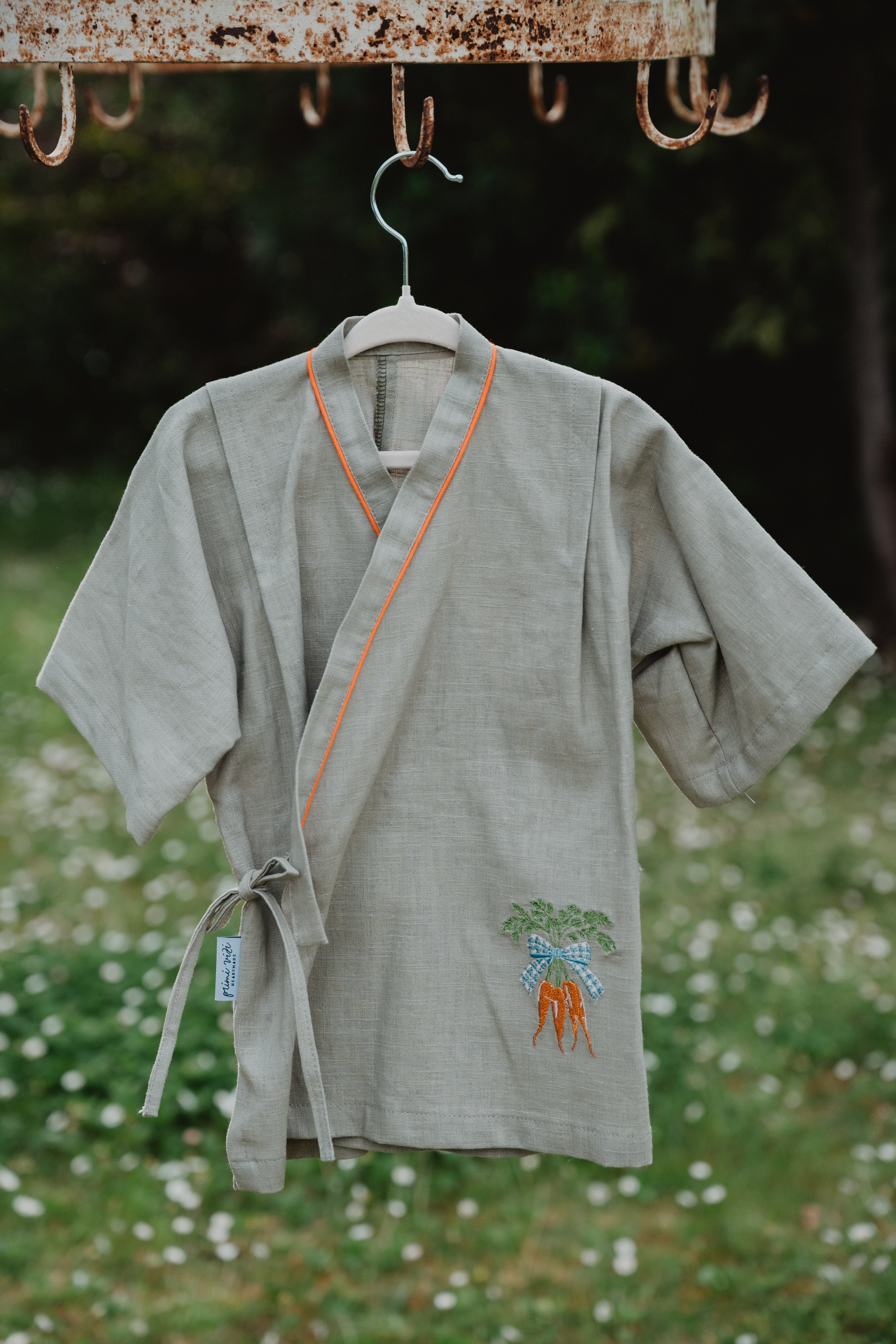 Camicia kimono in lino lavato verde oliva appesa, bordo arancione, ricamo carote